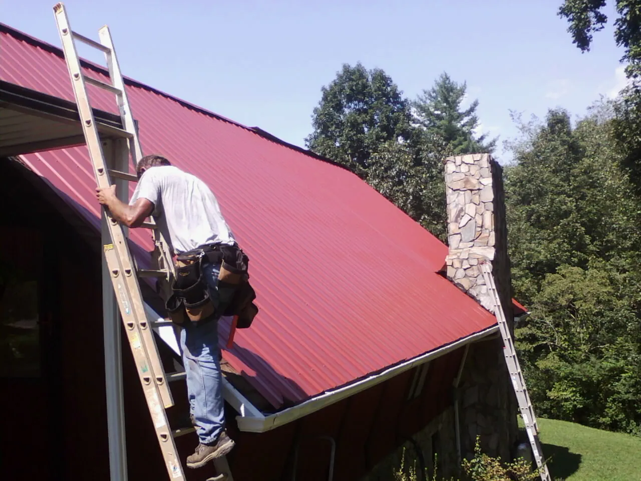 Roofer on a ladder beside a red metal roof and stone chimney — Soffit & Fascia Repair by Roofing Brothers in Naselle